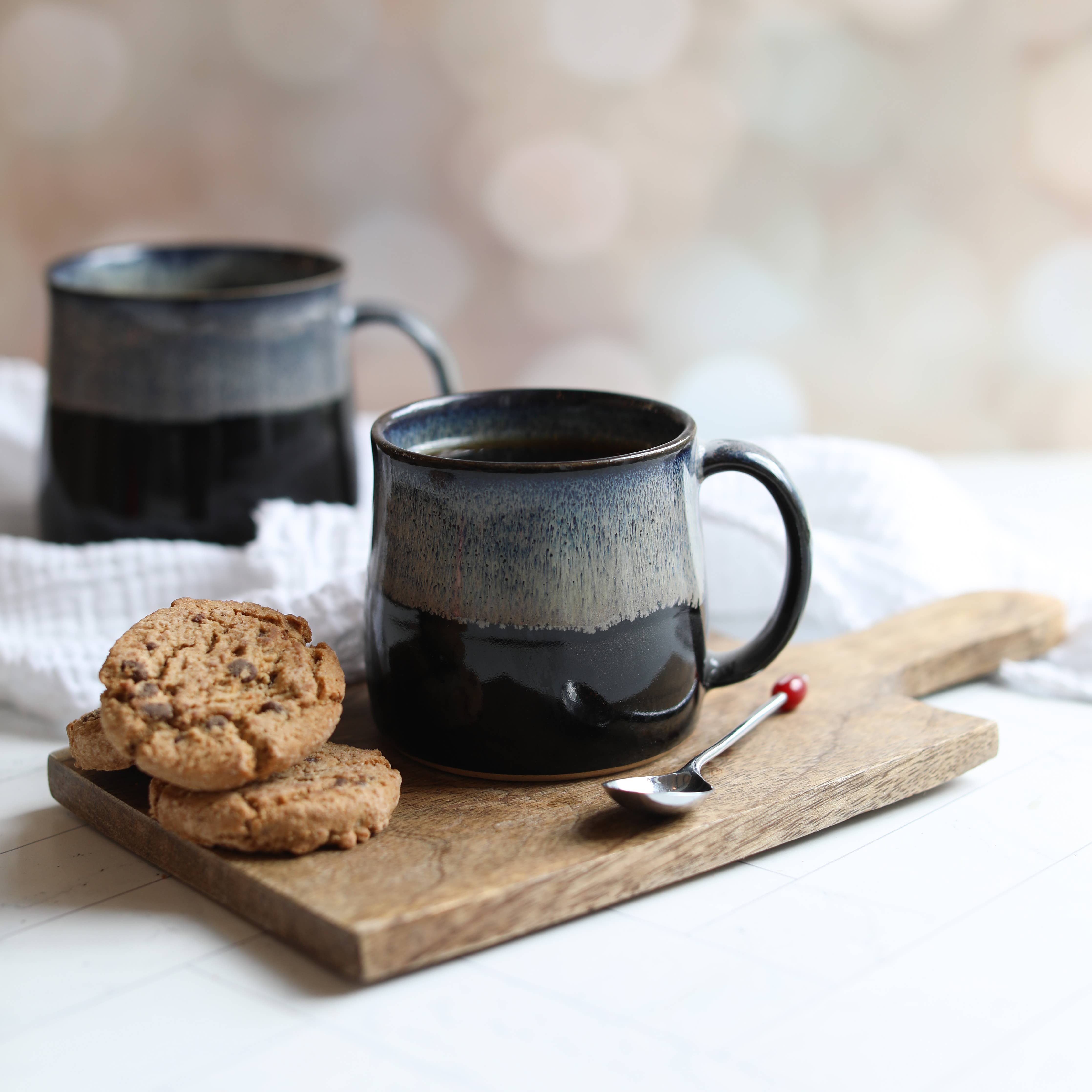 Two black ceramic mugs on a wooden board with cookies and a spoon.