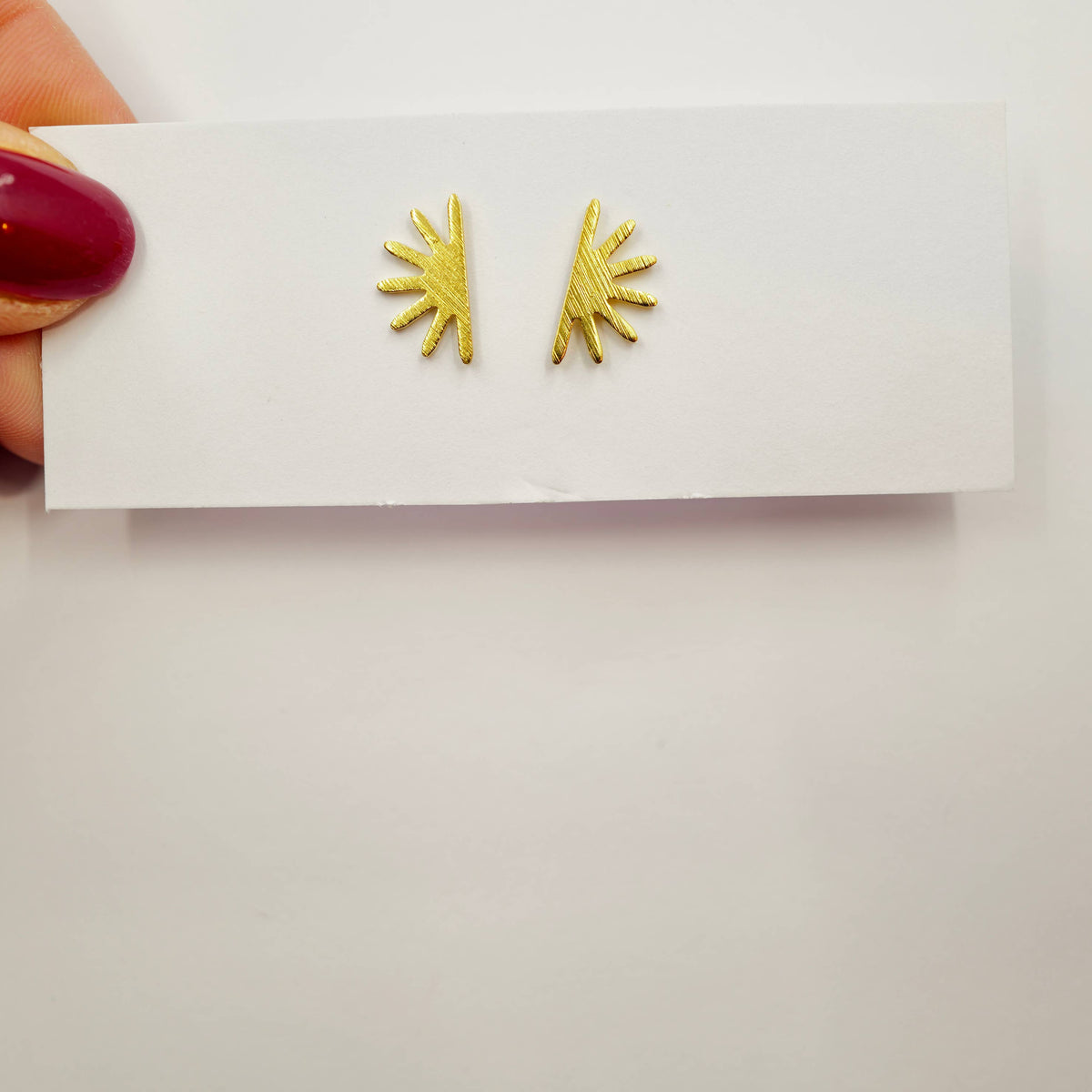 Gold sunburst earrings on a white card held by a hand with red nail polish.