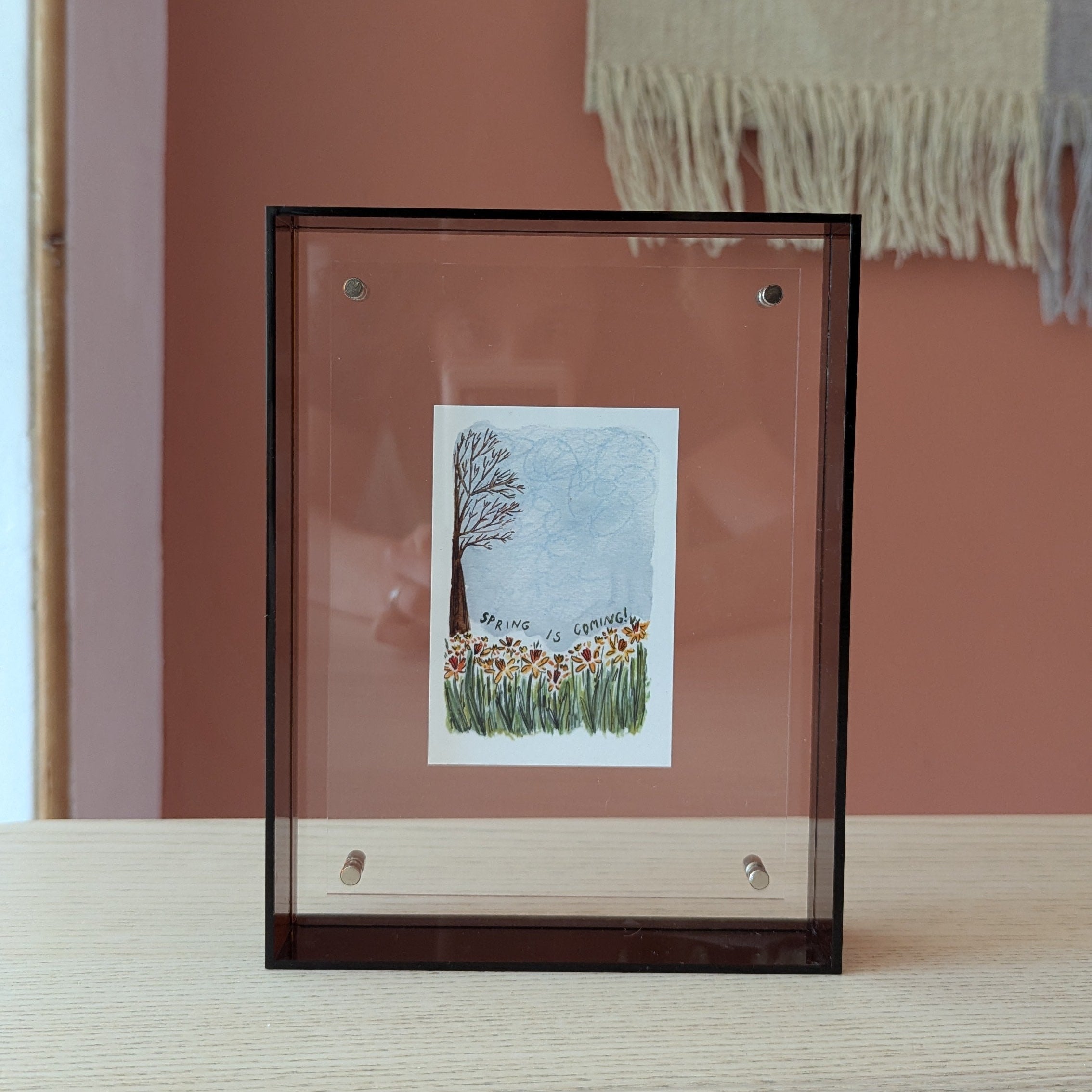 Framed artwork of a tree and flowers on a wooden surface with a pink wall in the background.