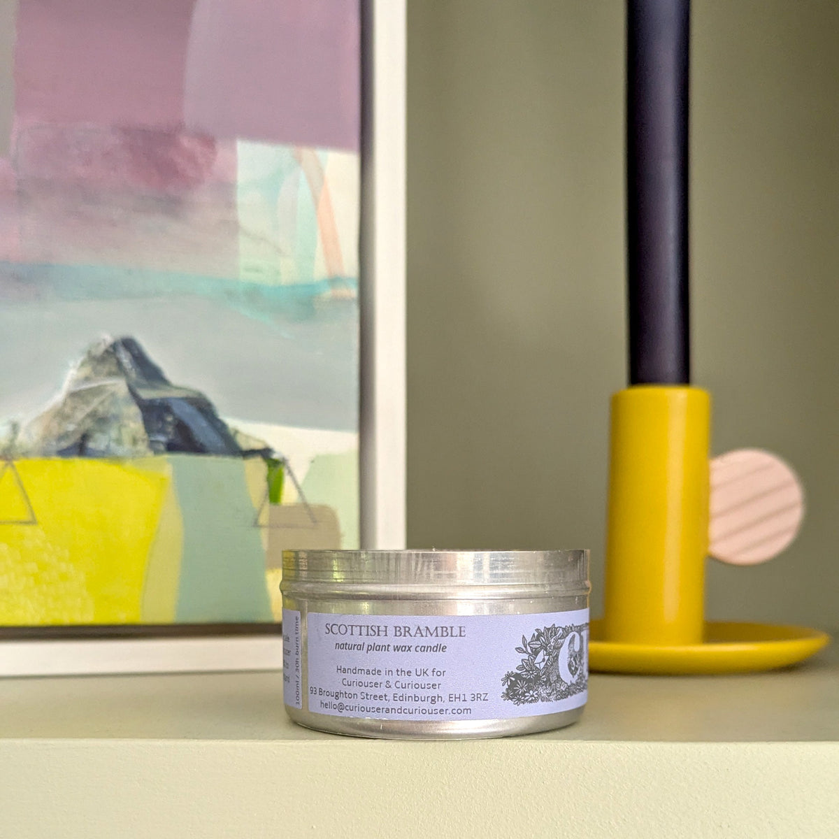 A small round tin of Scottish Bramble candle sits on a shelf in a styled interior setting with books, a colorful framed print, and a yellow candlestick holder in the background.