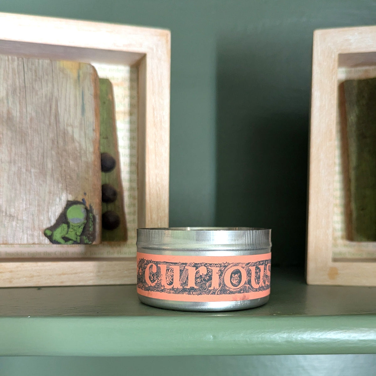 A small round tin candle with a silver lid and a peach-orange label reading 'CURIOUS' sits on a painted green shelf. Framed wall art and decorative objects are visible around it in a styled interior setting.