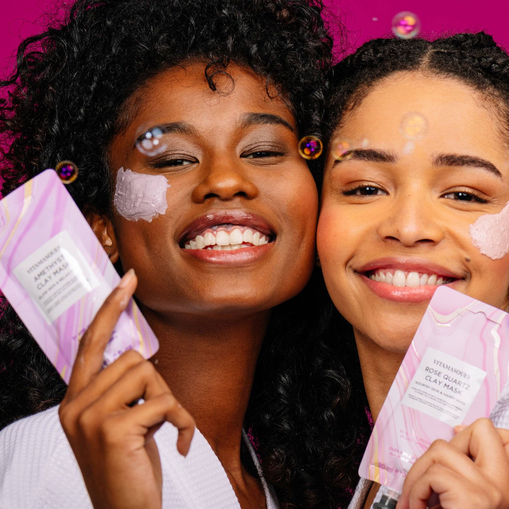 Two women with face masks on holding skincare products against a pink background