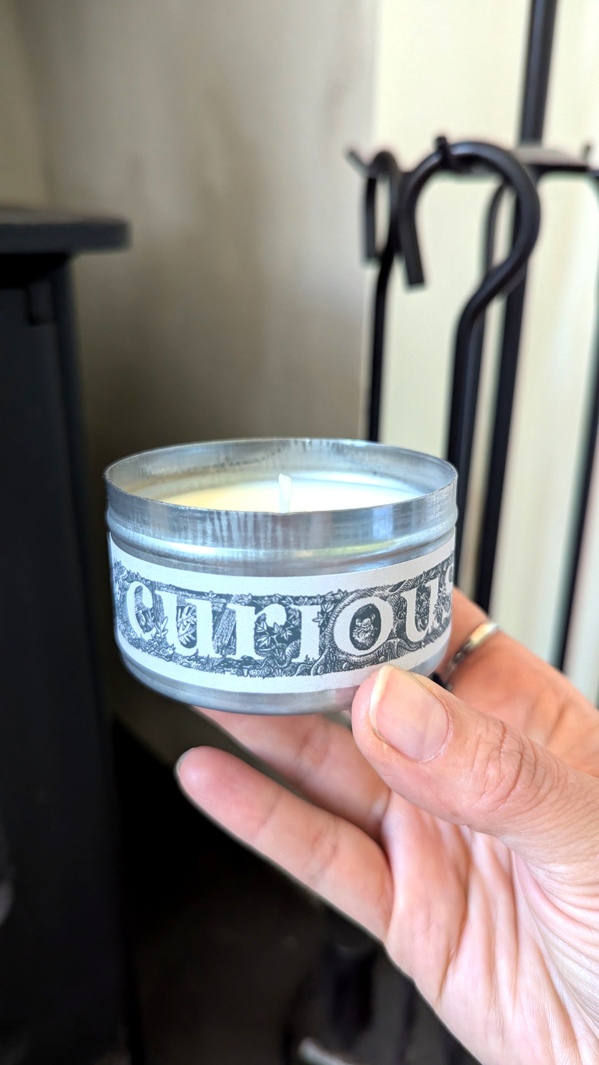 A hand holding a small round glass jar candle with a white wax fill and a white label that reads 'curious' in a decorative black font. The background shows a dark fireplace or hearth tool holder against a light wall.